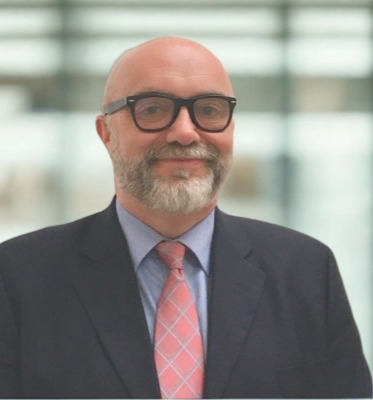 A white man with dark glasses smiles at the camera. He has a beard and is wearing a dark suit with a blue tie and a salmon pink tie  
